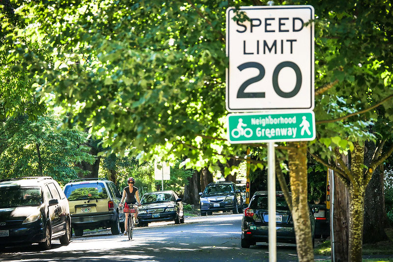 Woman riding bike on street with car parking and 20 mph speed limit and "Neighborhood Greenway" signs.